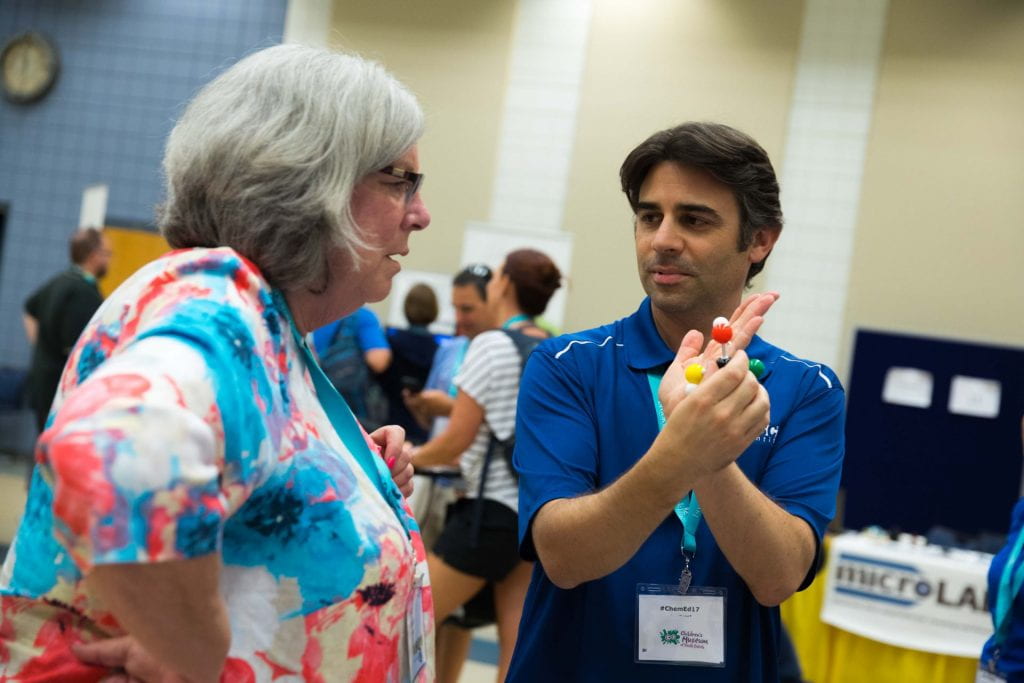 a man in a blue shirt holding a molecular model explaining something to a women in an exhibit hall