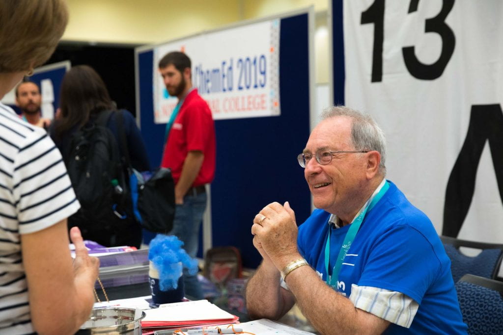 Lee Brubacher sitting at a exhibit hall table smiling and talking to people