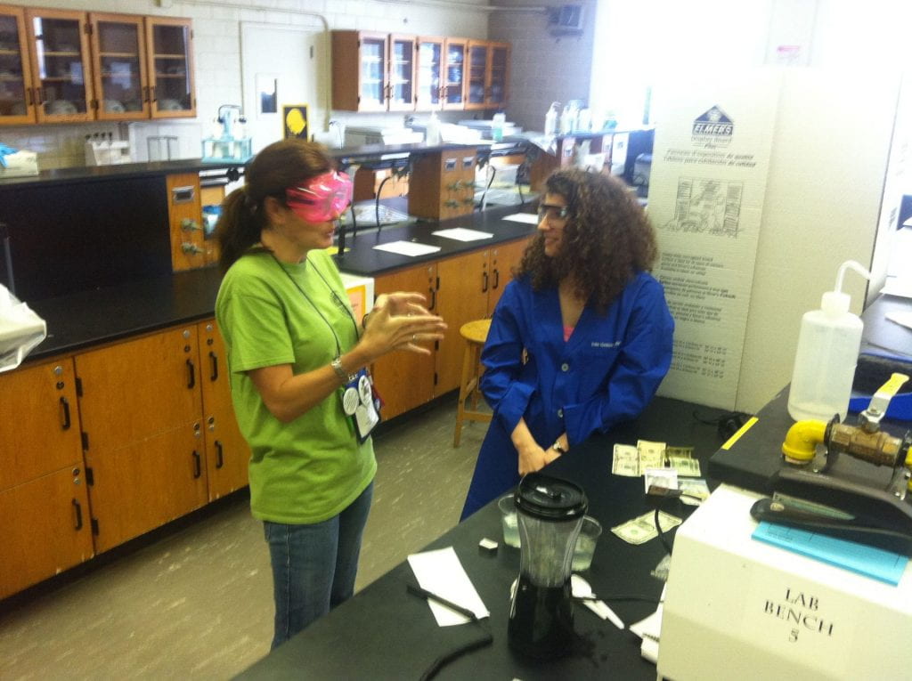 A women explaining a concept to another women in a lab setting, both wearing colourful goggles