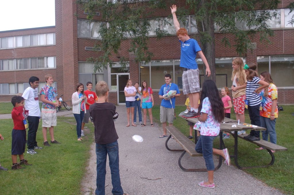 a boy standing on a picnic table surrounded by kids watching him drop a white paper pie plate