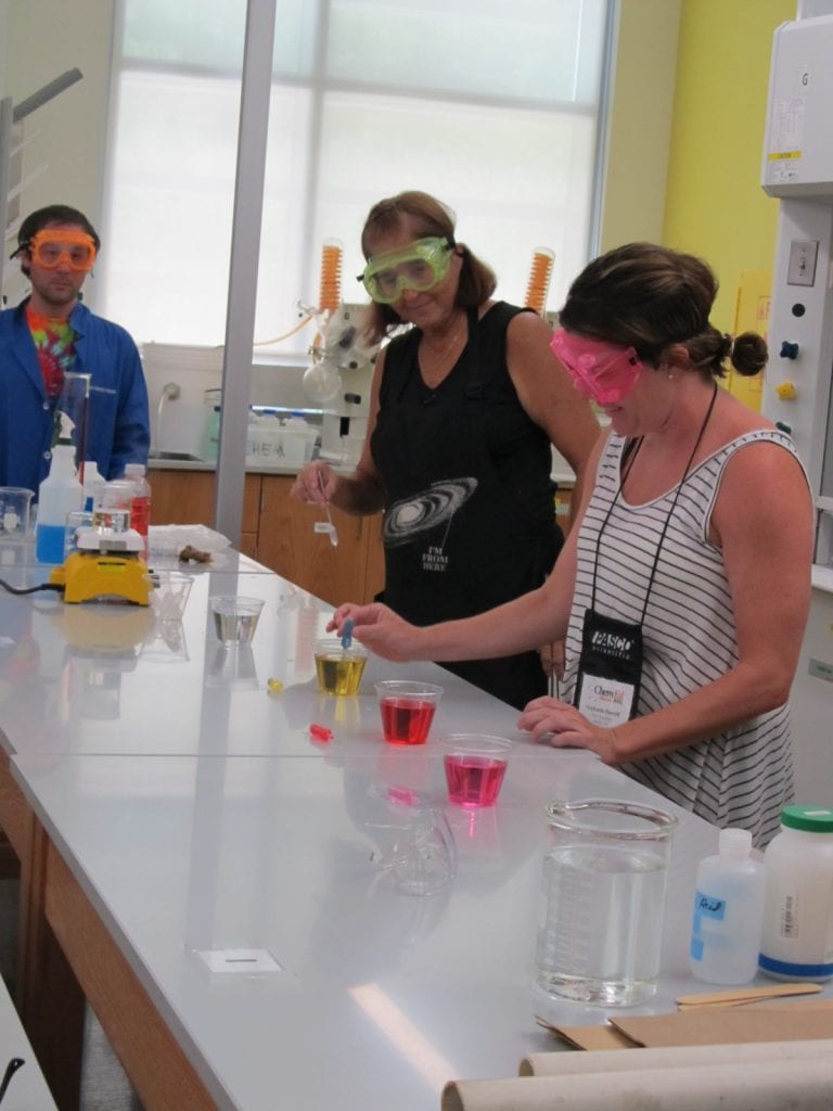 Peggy Sconzo in a lab setting with several plastic glasses holding colourful solutions instructing a workshop attendee with an eye dropper