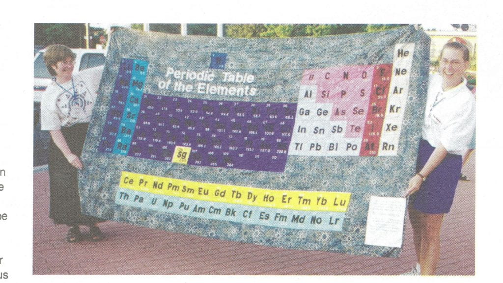 Two women holding a colourful periodic table quilt