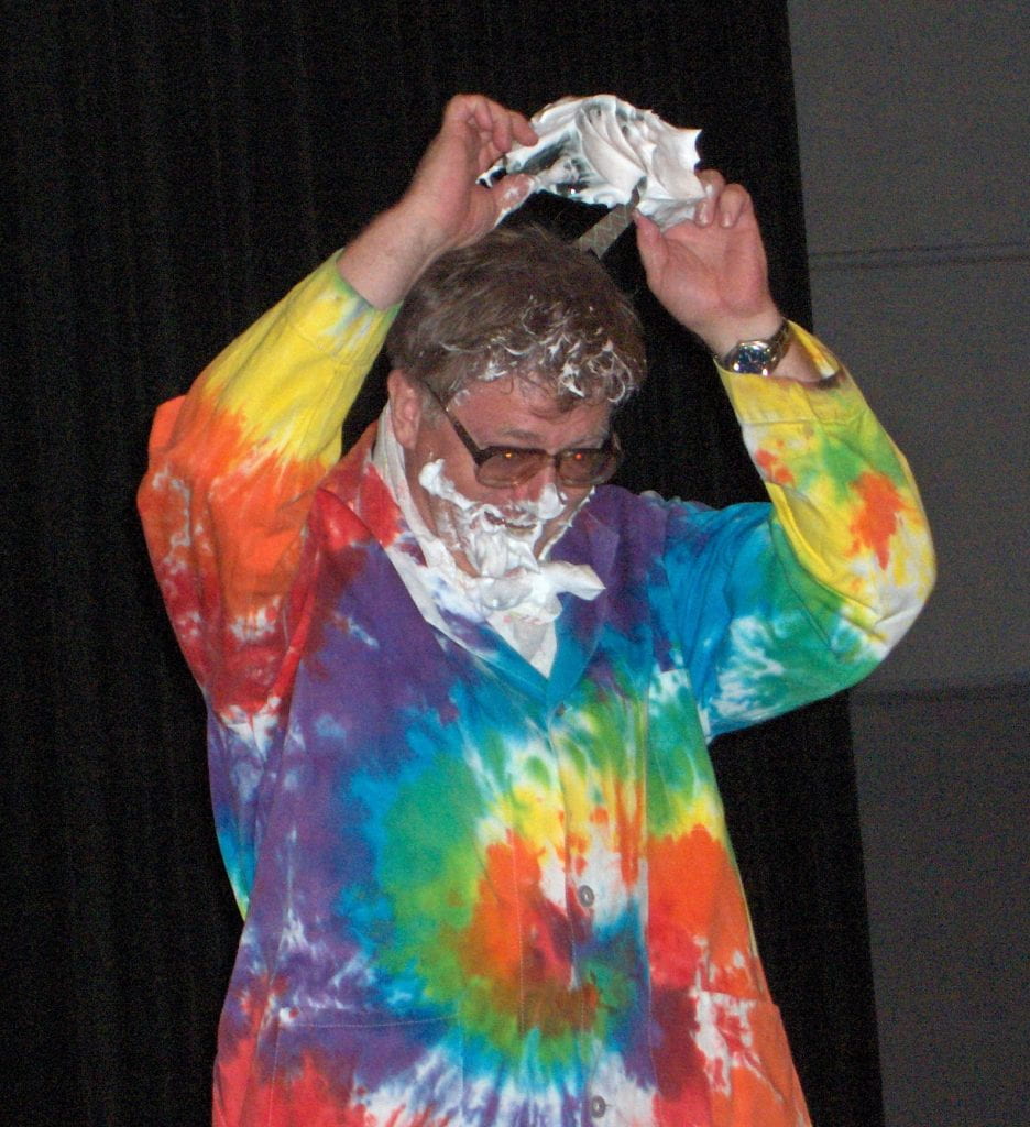 Andy Cherkas in a colourful tie dyed labcoat is pulling off goggles covered with white pie cream showing his eye are has been protected